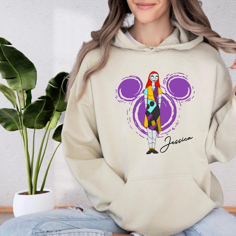 A woman wearing a beige hoodie with a colorful graphic of a character from the movie "Sneaky Nancy" printed on it.
