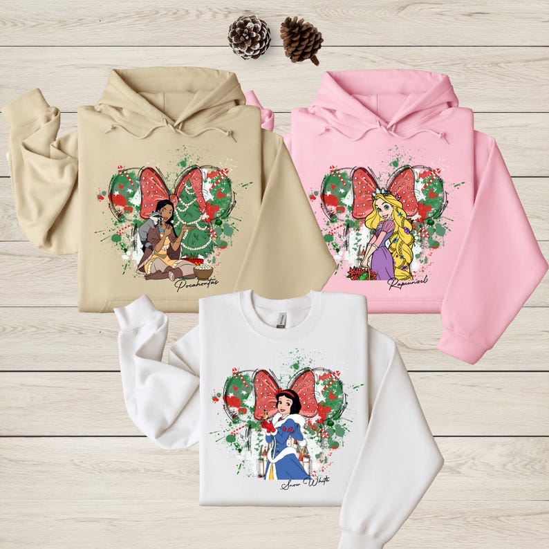 Three colorful hoodies with illustrations of Disney princesses are displayed on a wooden surface.