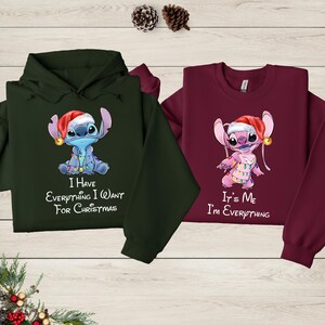two sweatshirts with a cartoon character from the Disney movie Lilo and Stitch. The sweatshirt on the left is green and the one on the right is burgundy. Both sweatshirts have a Christmas theme, with the character wearing a Santa hat and the text "I have everything I want for Christmas" printed on them.
