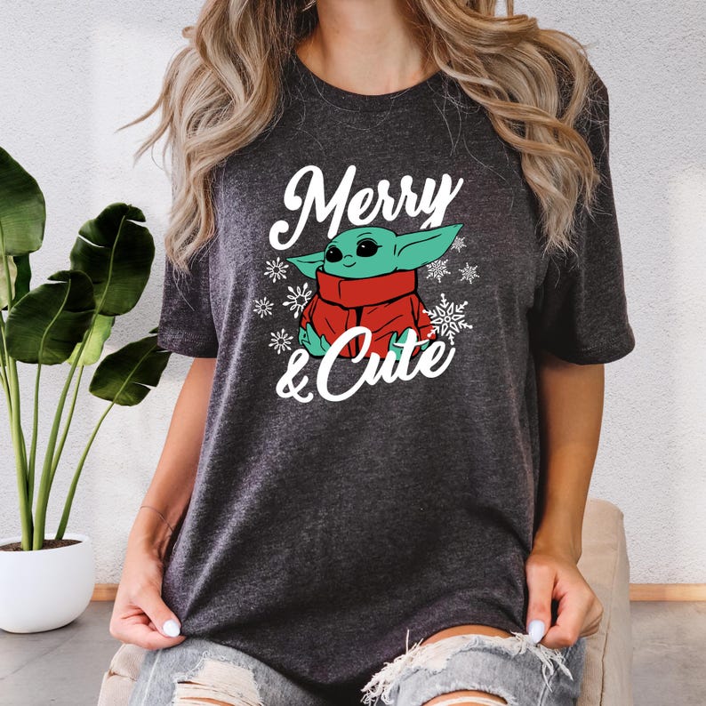 A woman wearing a gray t-shirt with a graphic design featuring a cute, cartoon-like character and the text "Merry & Cute".