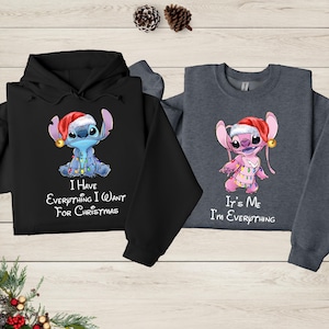 two sweatshirts with a cartoon character from the Disney movie Lilo and Stitch. The sweatshirt on the left is black and the one on the right is gray. Both sweatshirts have a humorous message printed on them, which reads "I have everything I want for Christmas" and "It's me, I'm everything".