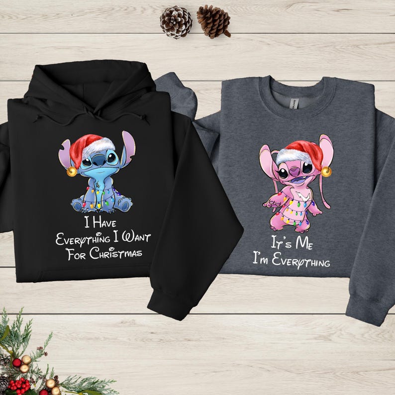 two sweatshirts with a cartoon character from the Disney movie Lilo and Stitch. The sweatshirt on the left is black and the one on the right is gray. Both sweatshirts have a humorous message printed on them, which reads "I have everything I want for Christmas" and "It's me, I'm everything".