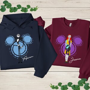 two sweatshirts with cartoon characters from the movie " jack Skellington and Sally Ann". The sweatshirt on the left is a navy blue hoodie with a skeleton and a mouse, while the sweatshirt on the right is a burgundy long-sleeved shirt with a girl and a mouse.