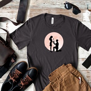 May include: A dark gray t-shirt with a graphic of a couple silhouetted against a pink moon. The man is kneeling and holding a bouquet of flowers, while the woman is standing and smiling.