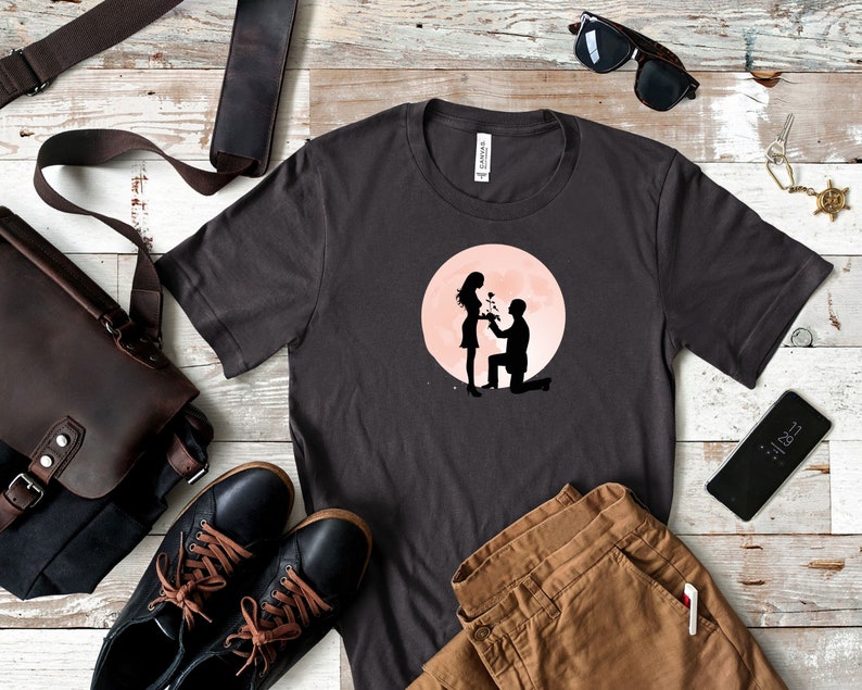 May include: A dark gray t-shirt with a graphic of a couple silhouetted against a pink moon. The man is kneeling and holding a bouquet of flowers, while the woman is standing and smiling.