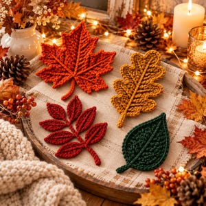 May include: A set of four crocheted leaf coasters in autumn colors, including a large orange maple leaf, a golden yellow oak leaf, a dark red compound leaf, and a deep green simple leaf, all arranged on a textured beige fabric surface.