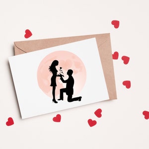 May include: A white card with a silhouette of a couple in love. The man is kneeling and holding a rose, while the woman is standing and smiling. The background is a pink circle, resembling a moon.