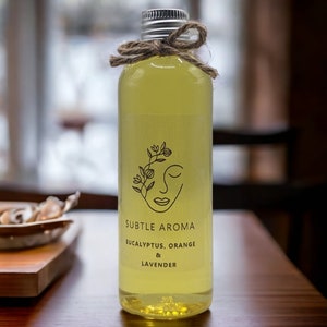May include: A clear glass bottle with a silver lid and a brown twine bow. The bottle contains a yellow liquid and has a label that reads "Subtle Aroma Eucalyptus, Orange & Lavender".
