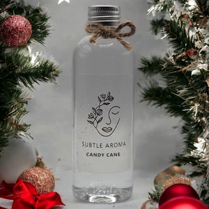 May include: A clear glass bottle with a silver lid and a brown twine bow. The bottle is filled with a clear liquid and has a white label that says "SUBTLE AROMA CANDY CANE". The bottle is surrounded by Christmas decorations, including red and gold baubles and a red bow.