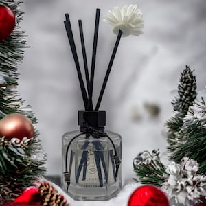 May include: A clear glass bottle diffuser with black reeds and a white flower on top. The bottle has a black label with the text "Subtle Aroma" and "Candy Cane". The diffuser is surrounded by Christmas decorations, including red and gold baubles, pine branches, and a red ribbon.