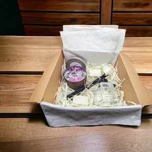May include: A brown cardboard box filled with a gift set of home fragrance products. The set includes a candle with dried flowers, a diffuser with reeds, a bar of soap, and incense sticks. The box is filled with white shredded paper.