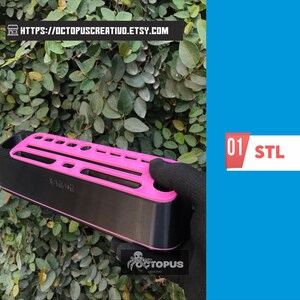 May include: A black and pink organizer with multiple slots, held by a gloved hand against a backdrop of green foliage. The organizer has the text "Octopus Creativo" and the text "01 STL".