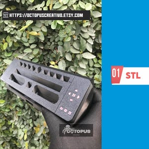 May include: A black 3D-printed organizer with various compartments and pink button details. The organizer is held against a backdrop of green foliage. The text "01 STL" is displayed on a white and red rectangle.
