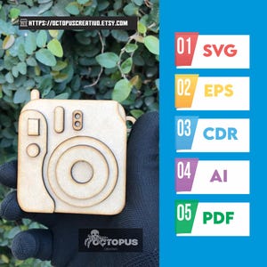 May include: A wooden camera-shaped craft held against a backdrop of green foliage. The camera design includes a lens, viewfinder, and other details. The image also features a blue background with numbered file format options: SVG, EPS, CDR, AI, and PDF.