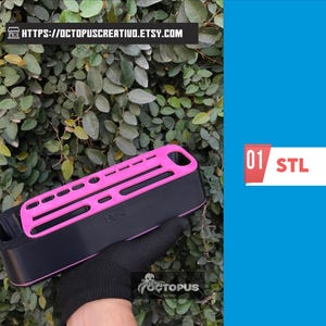 Może przedstawiać: Czarny i różowy prostokątny przedmiot z serią otworów i szczelin, trzymany przez dłoń w rękawicy. Obiekt ma tekst "01 STL" na białym i czerwonym prostokącie. Tło to zielona, liściasta roślina.