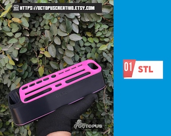 May include: A black and pink rectangular object with a series of holes and slots, held by a gloved hand. The object has the text "01 STL" on a white and red rectangle. The background is a green leafy plant.
