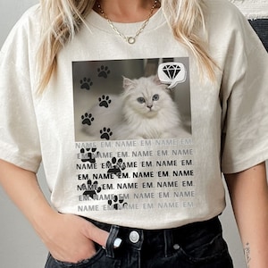 Puede incluir: Una camiseta beige claro con un gato blanco que lleva un collar de diamantes en una burbuja de diálogo. El gato está rodeado de huellas de patas negras. El texto "NAME EM. NAME EM. NAME EM. EM. NAME EM. NAME EM. NAME 'EM. NAME 'EM. NAME 'EM. EM, NAME EM. NAME EM. NAME EM. NAME EM. NAME EM. NAME EM. NAME EM. NAME EM" está impreso en la camiseta.
