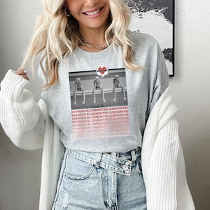Könnte beinhalten: Ein graues T-Shirt mit einem Schwarzweißbild von drei Skeletten, die auf einem Sims sitzen. Die Skelette tragen Hüte und haben eine Sprechblase über sich, auf der "REUNIONS" steht. Der Text "DIABOLICAL.DEMENTED.SUBHUMAN." wird mehrmals auf dem T-Shirt wiederholt.