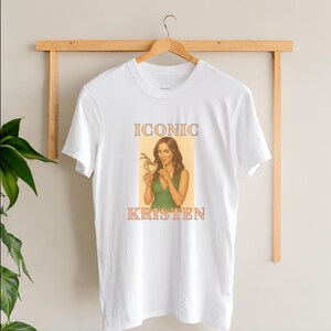 May include: White t-shirt featuring a retro-style graphic of a woman holding a coffee cup, with the text "ICONIC KRISTEN" in a peach color. The woman is wearing a green dress. The shirt is hanging on a wooden hanger.