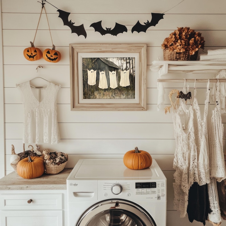 May include: A white laundry room with a washing machine, a clothes rack with white lace tops, and a framed picture of ghosts hanging on the wall. There are also pumpkins and a black bat garland on the wall.