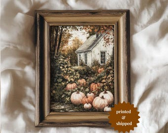 May include: A framed print featuring a quaint white cottage surrounded by pumpkins and autumn foliage. The artwork is set in a gold-toned frame. The print has a warm, inviting color palette, perfect for fall decor.