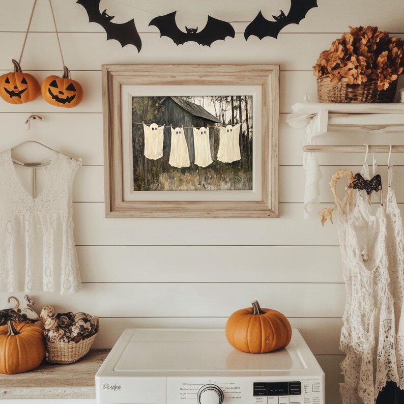 May include: A framed print of a vintage farmhouse with four white ghosts hanging on a clothesline. The farmhouse is painted in shades of gray and brown. The ghosts are cut out of white paper and are hanging from a string. The print is surrounded by a wooden frame. There are two pumpkins in the foreground, one orange and one white.