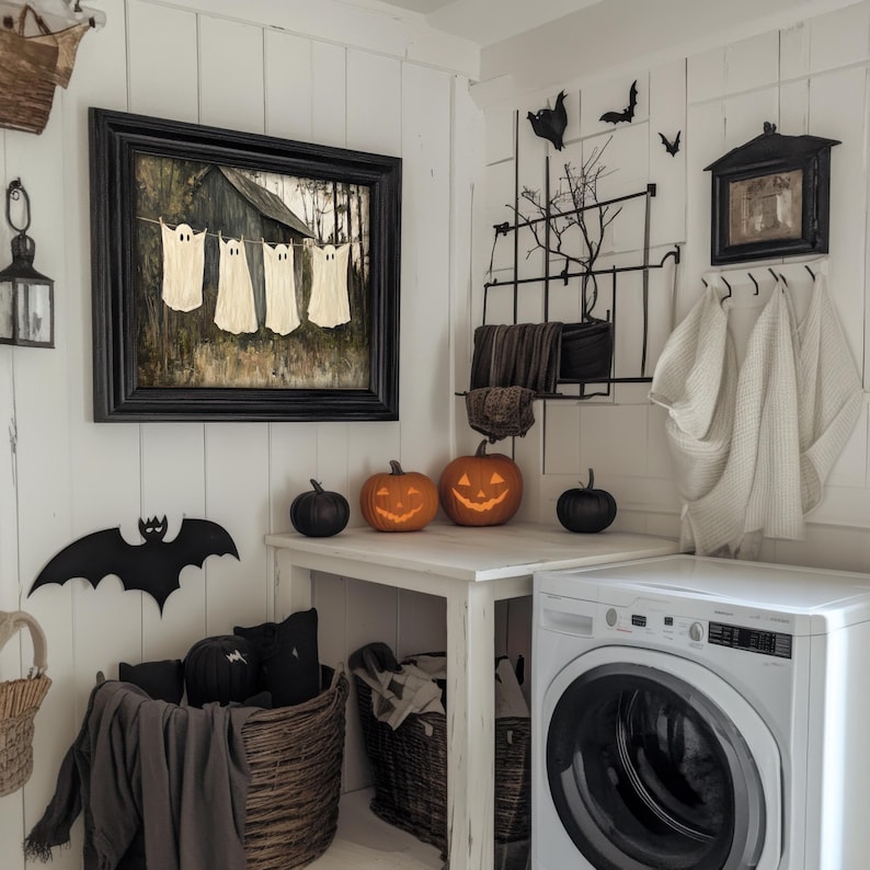 May include: A white laundry room with a black framed picture of ghosts hanging on the wall. There are carved pumpkins, a black bat, and a white washing machine in the room.