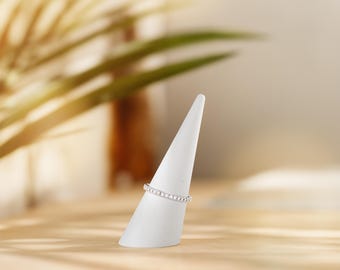 May include: A silver ring with a row of small, clear stones is displayed on a white, cone-shaped ring holder. The ring holder is on a light brown wooden surface. The background is blurred, with green foliage and soft lighting.