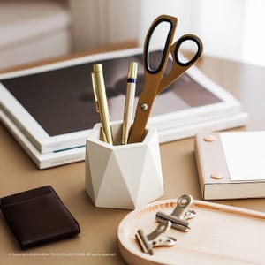 Op de afbeelding: Een witte geometrische pennenhouder met gouden pennen en een gouden schaar. Een kleine bruine portemonnee en een houten dienblad met metalen clips zijn ook zichtbaar. De items zijn gerangschikt op een lichtbruin oppervlak.