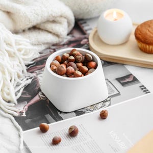 Könnte beinhalten: Eine weiße Schale gefüllt mit Haselnüssen steht auf einer bedruckten Oberfläche. Eine brennende Kerze und ein Muffin befinden sich im Hintergrund. Eine gemütliche, weiße Decke ist in der Nähe drapiert, was eine warme Atmosphäre schafft.