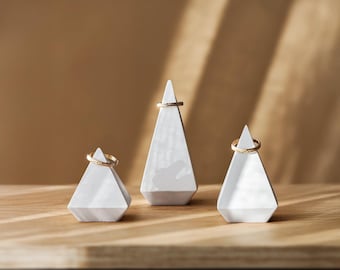 May include: Three geometric ring holders in a row. Each holder is a white, triangular prism with a gold ring on top. The holders sit on a wooden surface against a neutral background. The copyright notice at the bottom reads "Copyright 2024 KARA-TEKE COLLECTIONS. All rights reserved."