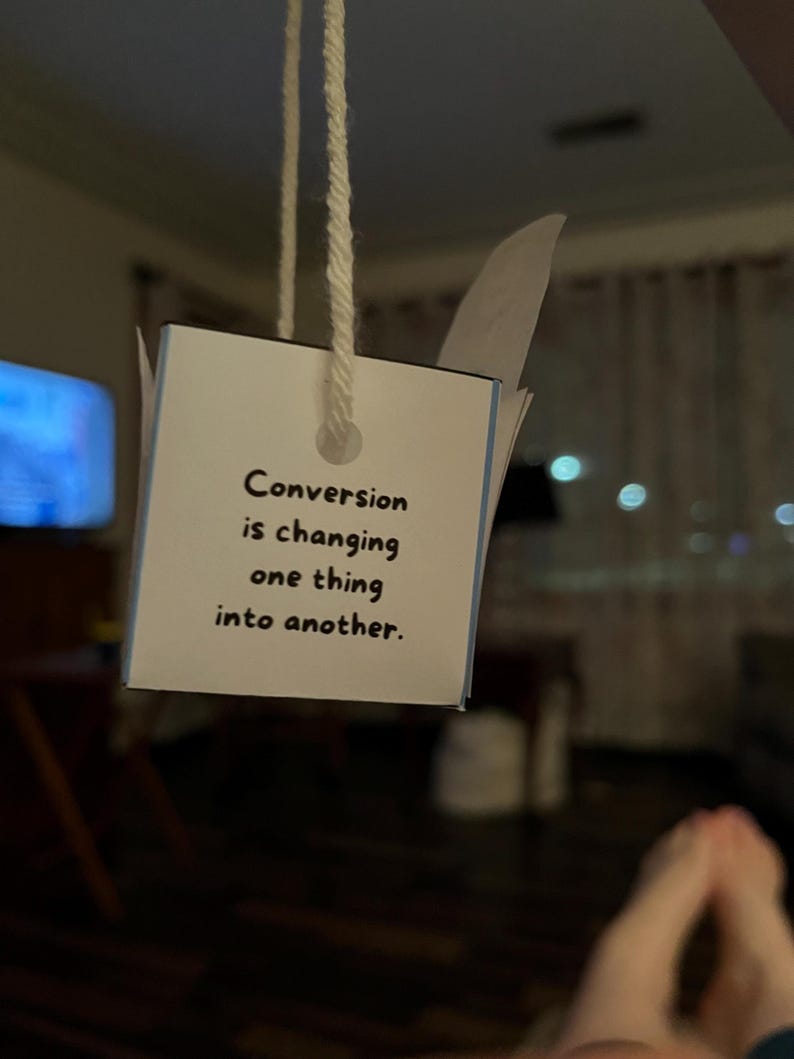 Peut inclure: Une petite carte carr&eacute;e avec le texte "Conversion is changing one thing into another" est suspendue par une ficelle blanche. La carte est blanche avec des accents bleus. L'arri&egrave;re-plan est flou, avec des indices d'une pi&egrave;ce et d'une t&eacute;l&eacute;vision.