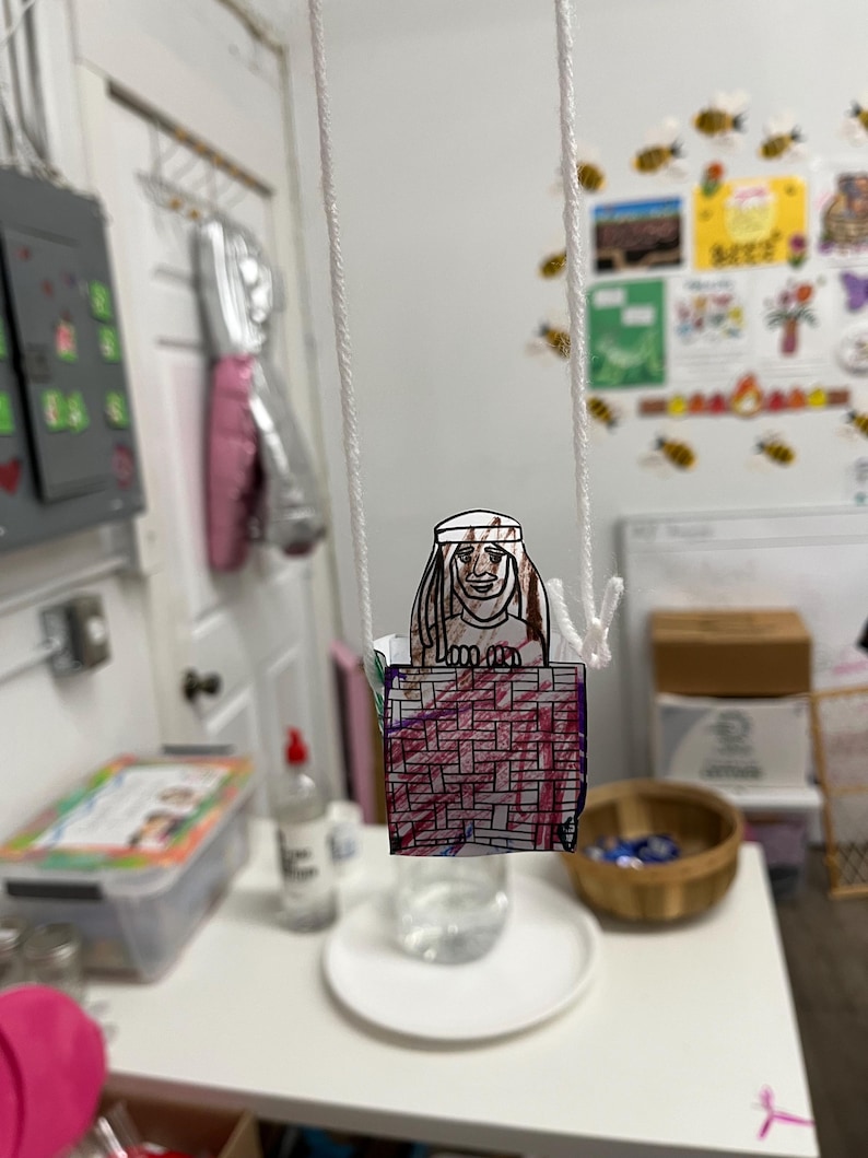 Peut inclure: Un artisanat en papier fait &agrave; la main repr&eacute;sentant une figure avec un couvre-chef, suspendue par une ficelle blanche. La figure est au-dessus d'une base rectangulaire avec un motif rouge et violet. L'arri&egrave;re-plan comprend une table blanche et divers objets.