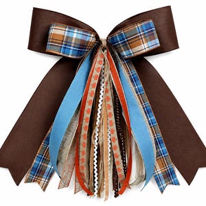 May include: A decorative bow featuring wide solid brown ribbon loops and long hanging streamers. The streamers include blue, orange, and brown plaid patterns, solid light blue, orange, and tan ribbons, and a thin ribbon with orange pumpkin prints.