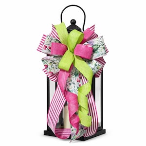 May include: Black lantern with a decorative bow. The bow features bright pink, lime green, and floral patterned ribbons. The lantern has a handle and a candle inside. A festive accent for home decor.