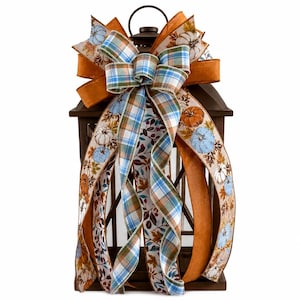 May include: A dark brown metal lantern adorned with a decorative multi-layered bow. The bow features ribbons in solid orange, blue and white plaid, and a cream-colored fabric printed with small blue and orange pumpkins and autumn leaves.