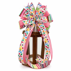 May include: A decorative bow with a dog theme, featuring colorful paw prints and bones on a white background. The bow is accented with pink and striped ribbons, and is placed on top of a wooden lantern.