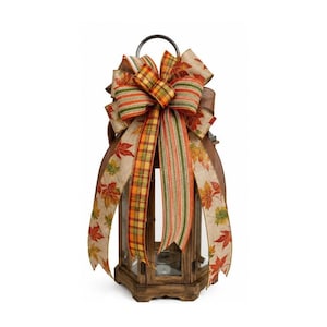 May include: A wooden lantern topped with a decorative bow featuring a mix of burlap ribbons. The ribbons display patterns of autumn leaves in red, orange, and green, alongside plaid and striped designs in matching fall colors with frayed edges.