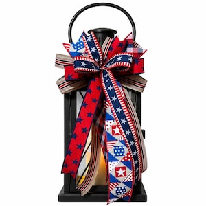 May include: A black metal lantern with a decorative patriotic bow. The bow features red, white, and blue ribbons with star and stripe patterns. The lantern has a handle and a warm light inside, perfect for home decor.