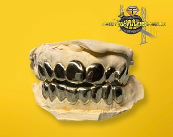 Puede incluir: Un juego de grillz dentales de color dorado, exhibido sobre un molde dental contra un fondo amarillo. Los grillz están muy pulidos y reflejan la luz, mostrando su acabado metálico. El logotipo "Sweet Tooth Gems Grillz" es visible en la esquina superior derecha.