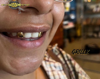 May include: Close-up of a person smiling, showing gold grillz on the upper teeth. The grillz are a shiny gold colour and appear custom-fitted. The image also includes the text "Sweet Tooth Gems & Grillz" and "Grillz".