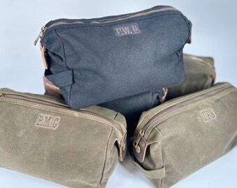 May include: Three toiletry bags made of canvas with leather accents. The bags are stacked on top of each other. The top bag is black with the initials "P.W.H." embroidered on the front. The middle bag is green with the initials "PMG" embroidered on the front. The bottom bag is green with the initials "TPB" embroidered on the front.