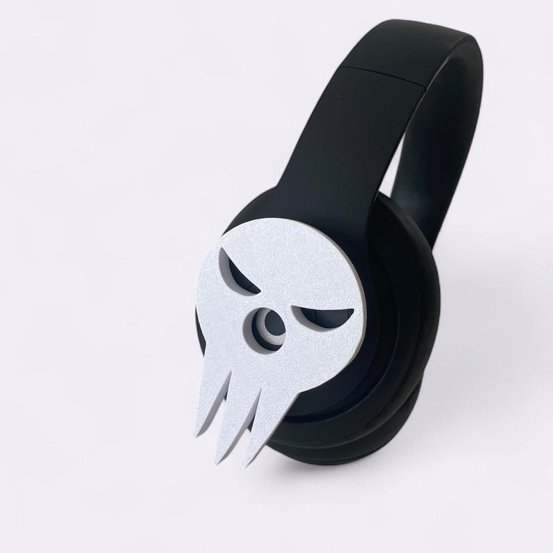 Peut inclure: Casque audio circum-auriculaire noir avec un motif de cr&acirc;ne argent&eacute; sur l'&eacute;couteur. Le cr&acirc;ne affiche une expression s&eacute;v&egrave;re avec des yeux obliques et une m&acirc;choire pointue. Le casque est pos&eacute; sur un fond blanc.