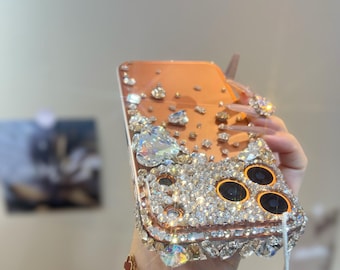 May include: A bedazzled phone case with a copper-colored back and a clear, gem-encrusted border. The case is adorned with various sizes of clear and iridescent crystals. The camera lenses are outlined in black.