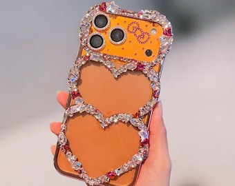 May include: A bedazzled phone case with a heart-shaped design. The case features a gold-colored background with a double heart outline made of clear and pink rhinestones. The top of the case has three camera lenses and a small graphic.