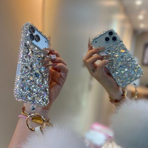 May include: Two silver phone cases are held up, displaying their sparkling, crystal-like embellishments. The cases are encrusted with numerous rhinestones, creating a glamorous design. The phone cases are a stylish accessory, perfect for adding sparkle.