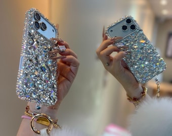Peut inclure: Une coque de smartphone recouverte de cristaux transparents à facettes qui reflètent la lumière. La coque présente une découpe pour triple objectif photo et est fixée à un anneau doré avec un pompon en fausse fourrure blanche.