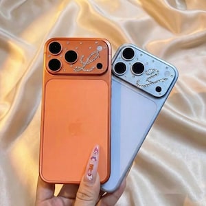 Puede incluir: Se muestran dos fundas para teléfono, una naranja y otra azul claro. La funda naranja tiene un diseño "Love" con pedrería y el logotipo de Apple. La funda azul claro también tiene un diseño "Love" con pedrería. Ambas fundas tienen un módulo de cámara con tres lentes negras.