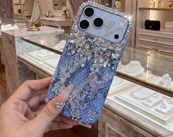 May include: A bedazzled phone case with a blue and silver design. The case is covered in sparkling rhinestones and crystals, with a gradient effect from silver to blue. The camera area is outlined with crystals.
