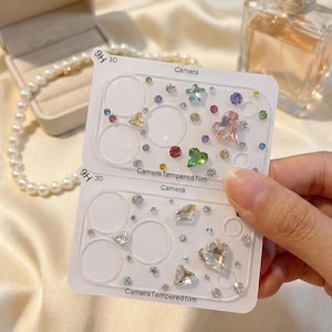 以下が含まれることがあります： 装飾的な宝石が付いた透明な携帯電話のカメラレンズプロテクター。「9H 3D」と「Camera Tempered film」の文字が入っています。宝石はさまざまな色と形をしています。